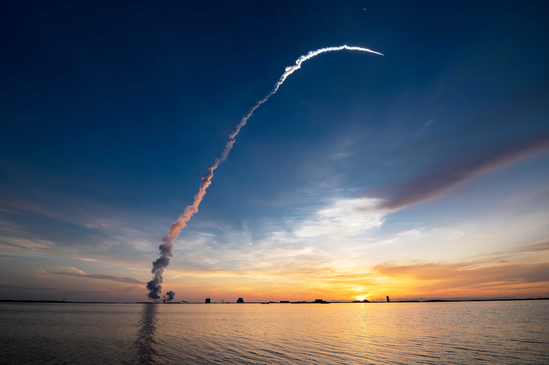 A rocket launches leaving behind a streak of smoke with an orangish sky in the background.