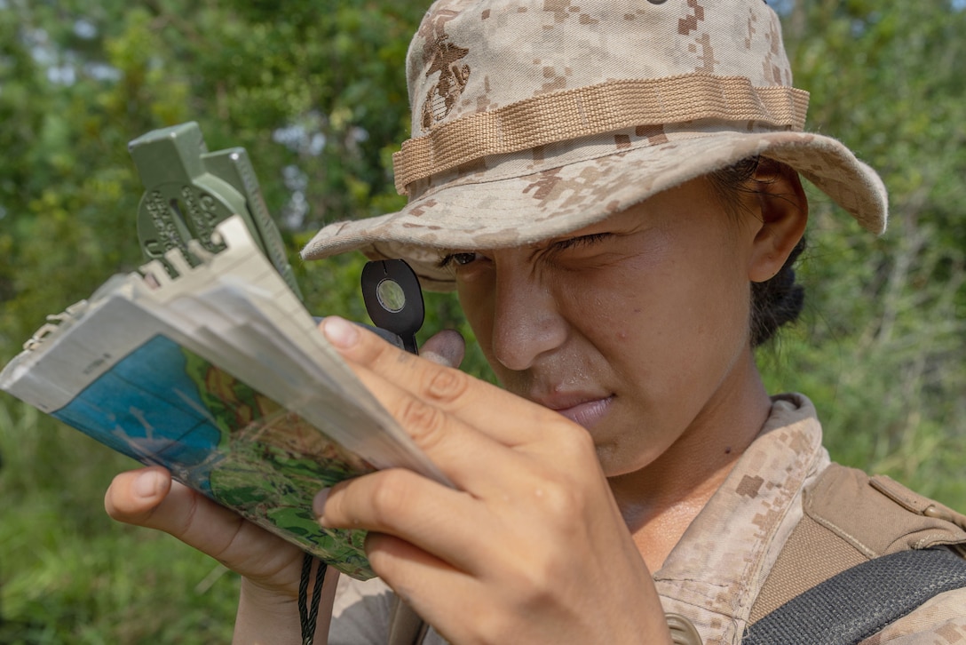 A close-up of a Marine Corps recruit looking through a magnifying glass at a piece of folded paper with trees in the background.