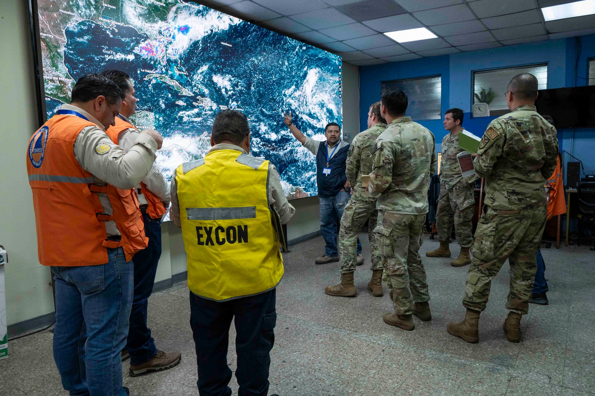 A photo of a group of people listening as a person points to a weather map.