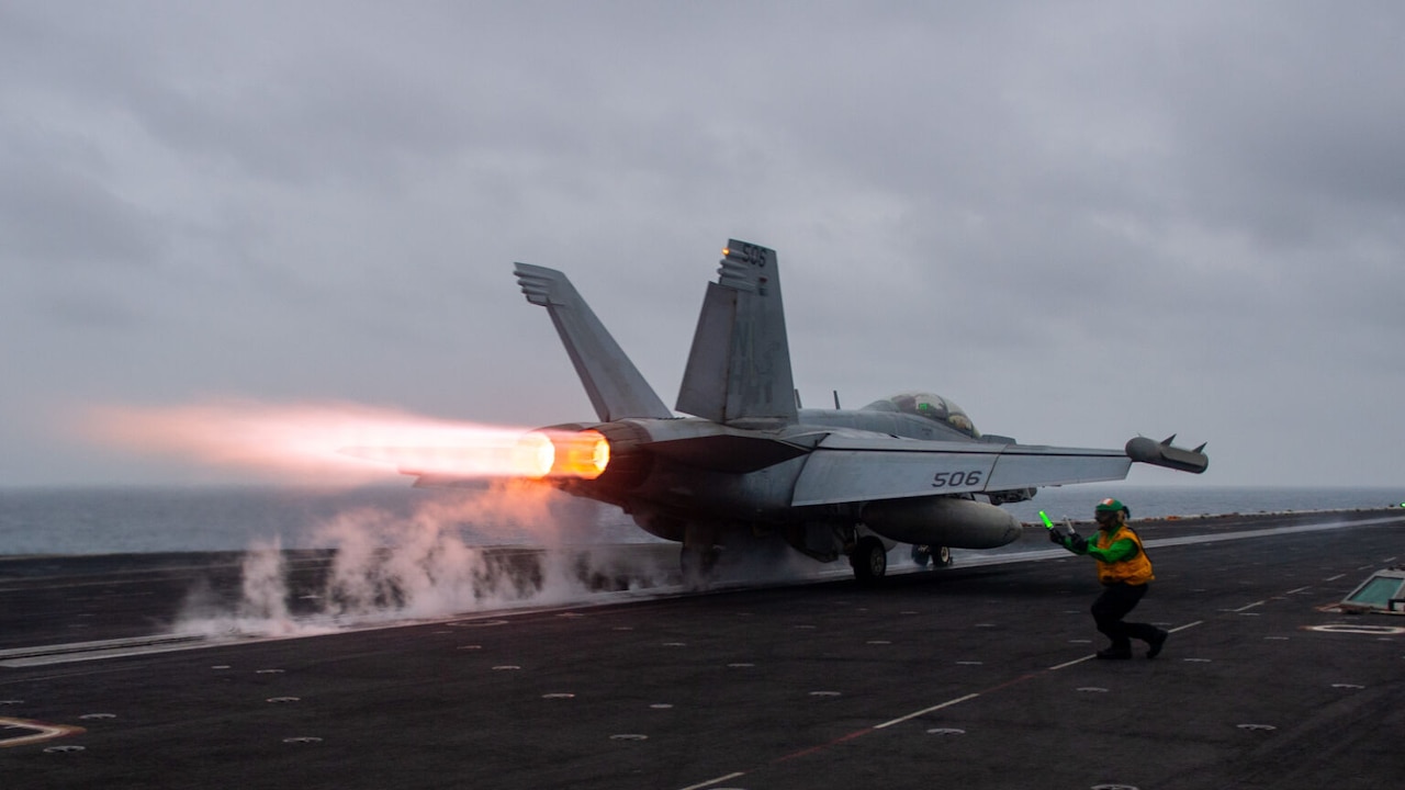 A service member kneels as a jet lands on an aircraft carrier.