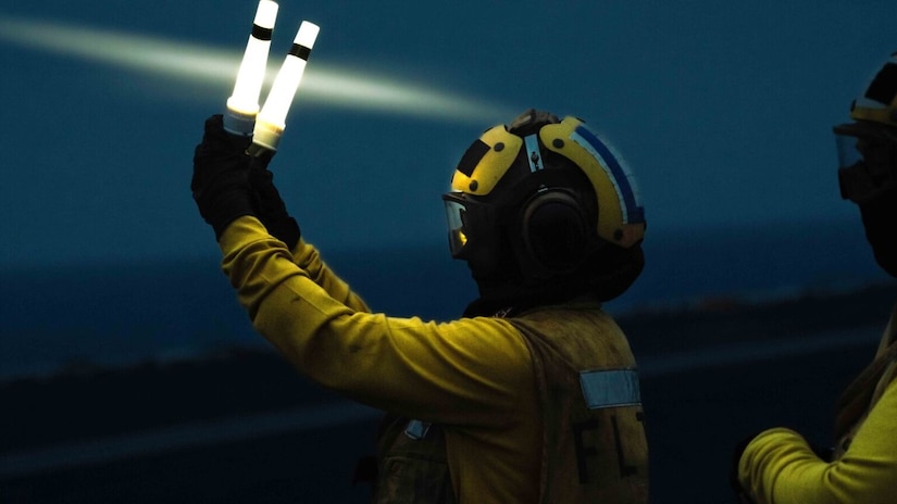 A service member holds lighted wands to wave an aircraft into position.