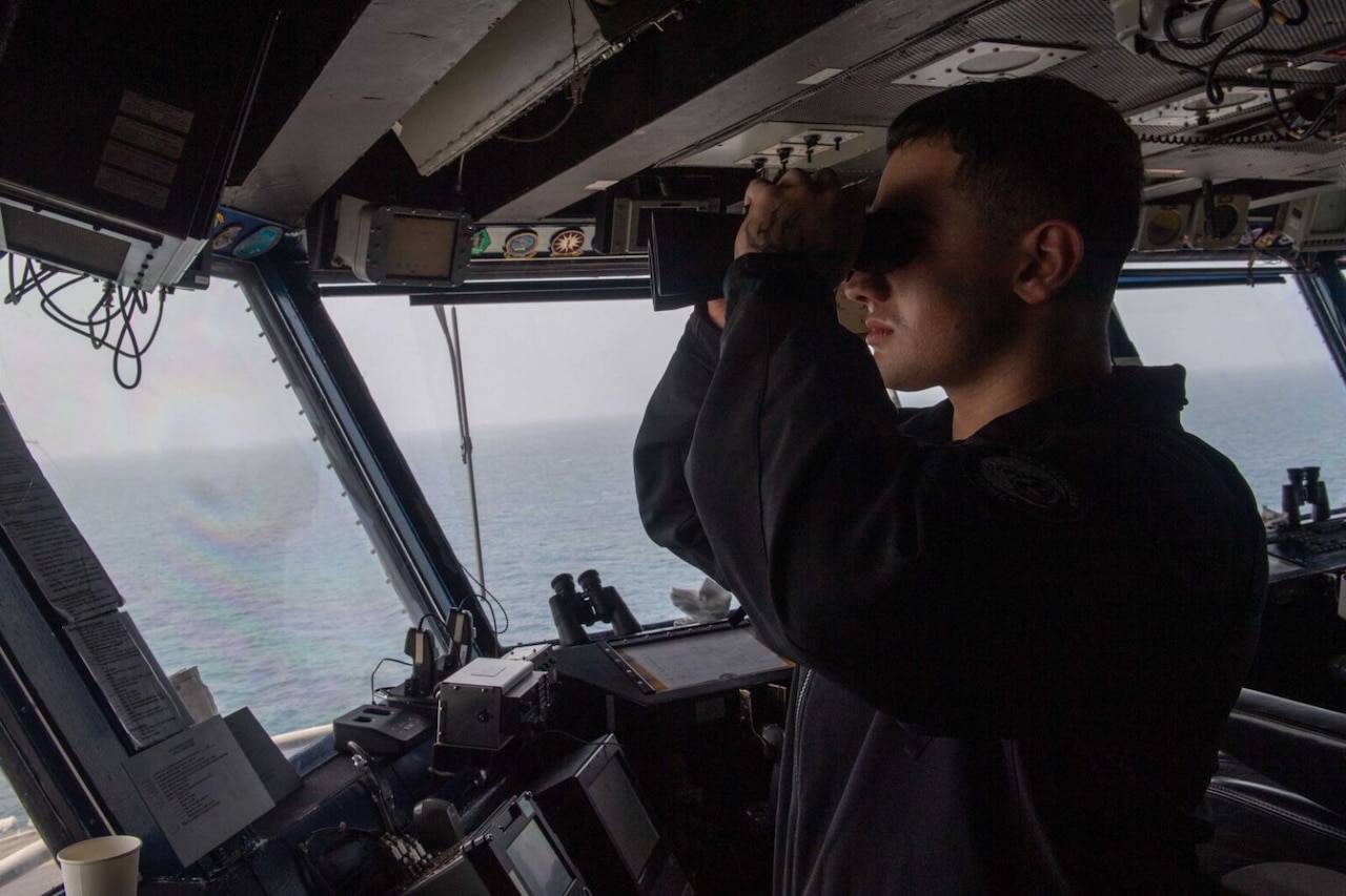 A service member looks through binoculars aboard a ship.