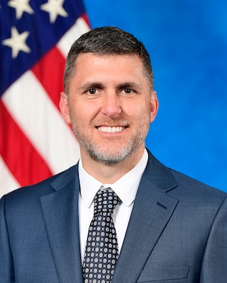 Dr. Jon T. Kosloski, a pale-skinned man with brown eyes, short salt and pepper hair and a short greying beard smiles broadly at the camera as he poses for an official photo in front of a blue background and American flag.