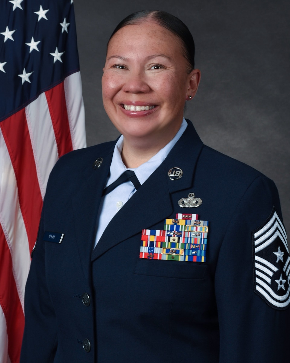 An Air Force command chief master sergeant in dress blues looks forward in front of an American flag and a grey background.