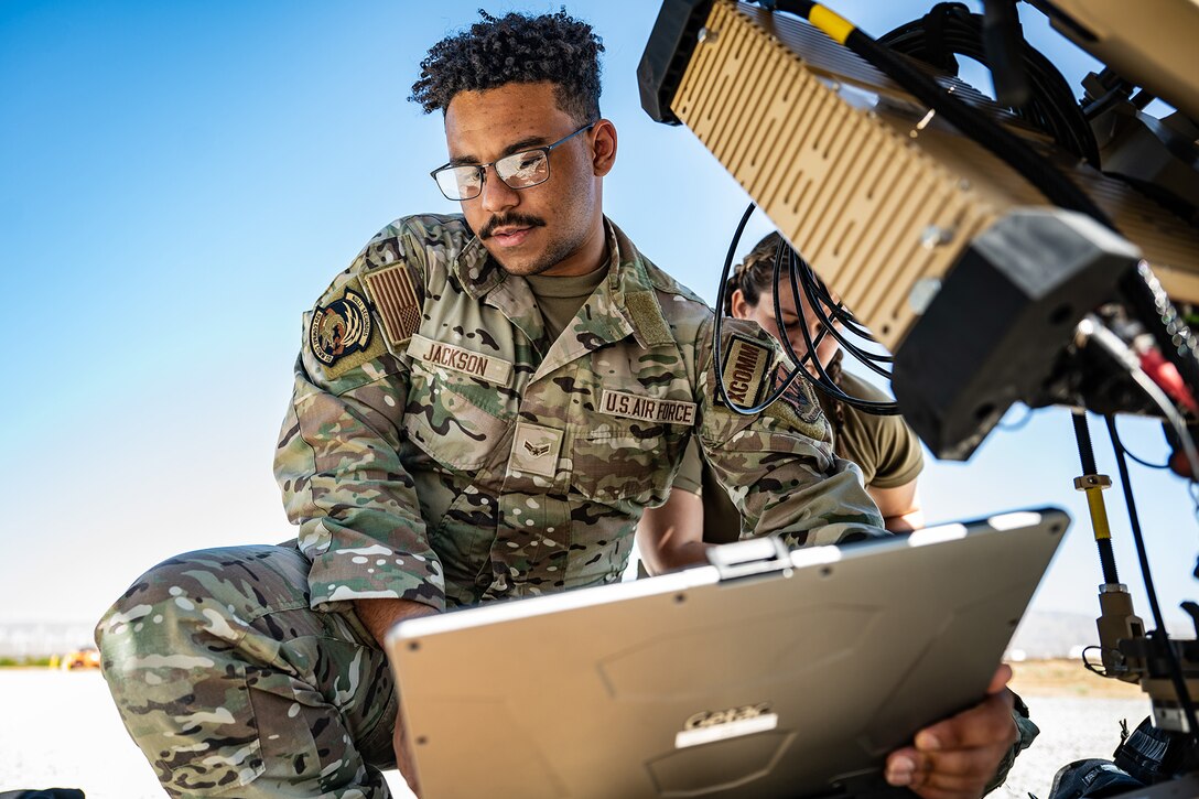 An airman sits in front of a laptop and other equipment during a clear and sunny day.