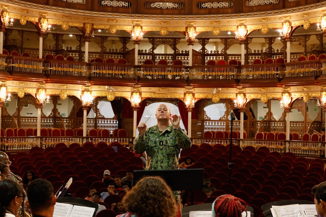 A Navy officer stands in front of a group of musicians holding a small conductor’s wand in a large opera house.