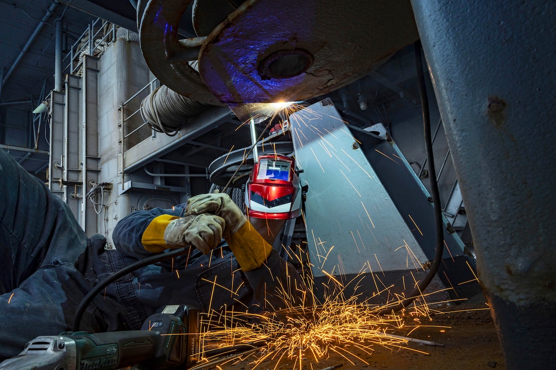 A sailor wearing a safety helmet and gear lies on the floor and uses a welding tool creating large orange sparks.