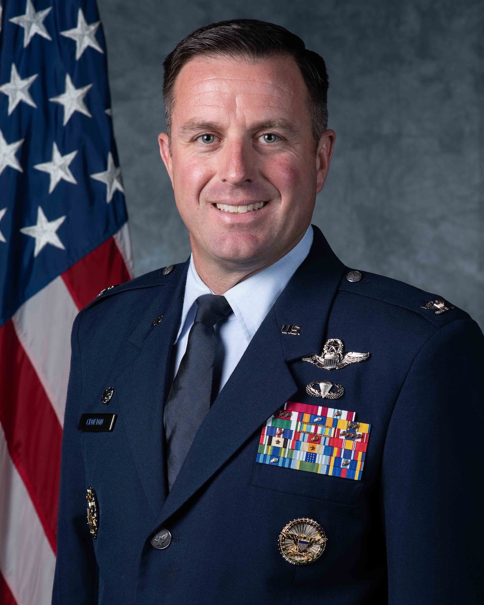 An Air Force commander in dress blues looks forward in front of an American flag and a grey background.