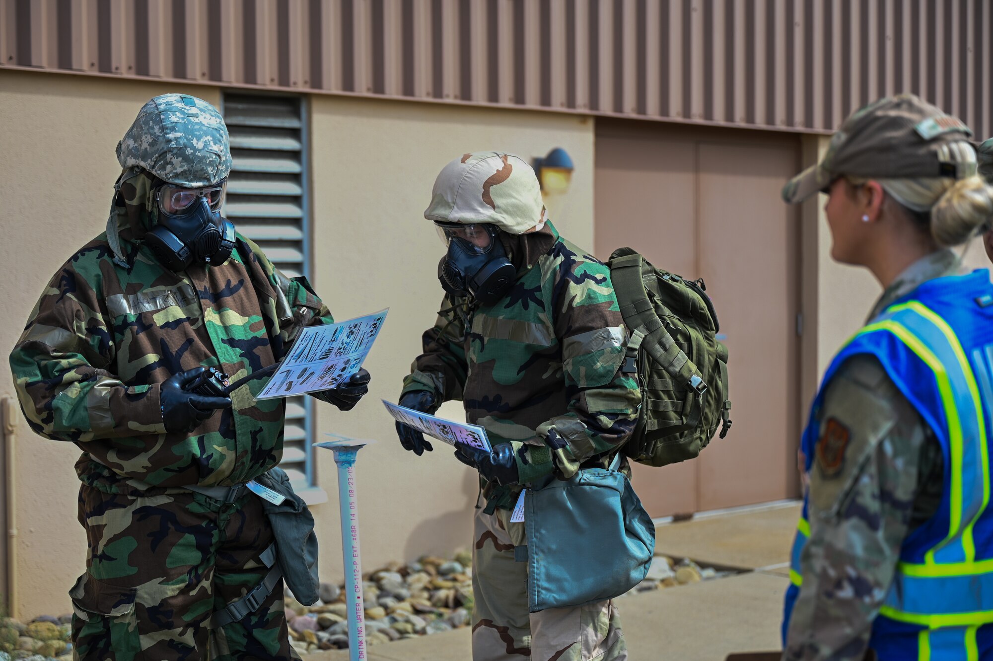Two Airmen wear HAZMAT suits while performing a check list while another Airman grades their performance.