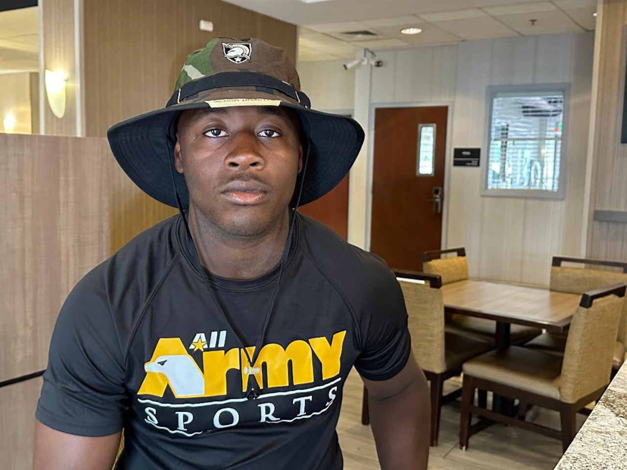 A man sits in a room and poses for a photo wearing a bush hat.