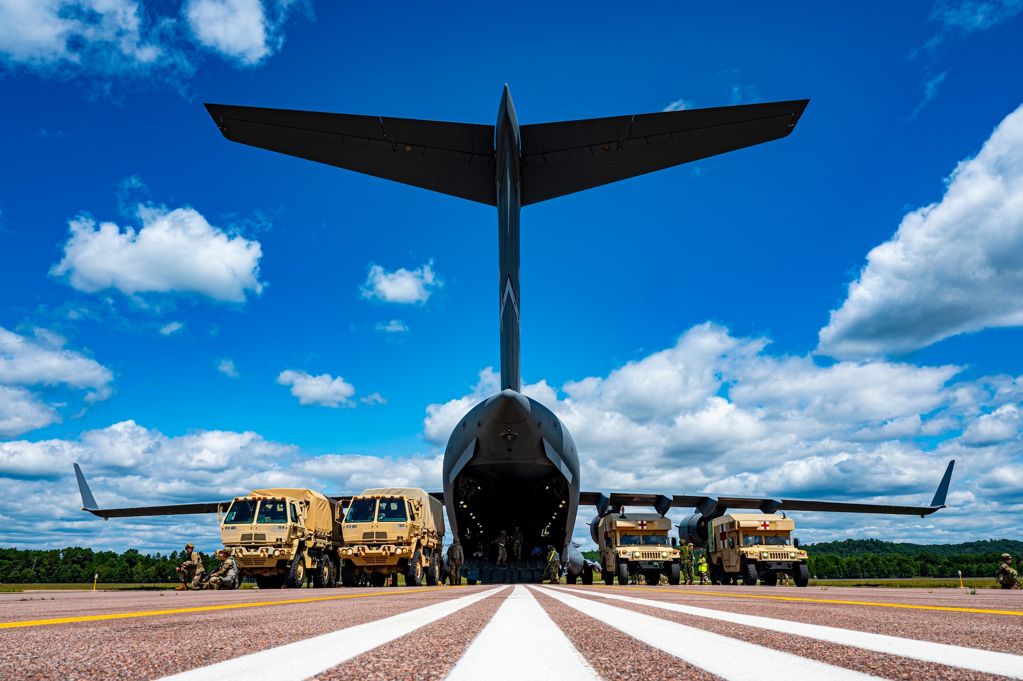 A fleet of Large Medium Tactical Vehicles align with the back of a C-17 Globemaster III on the Fort McCoy Sparta Airfield