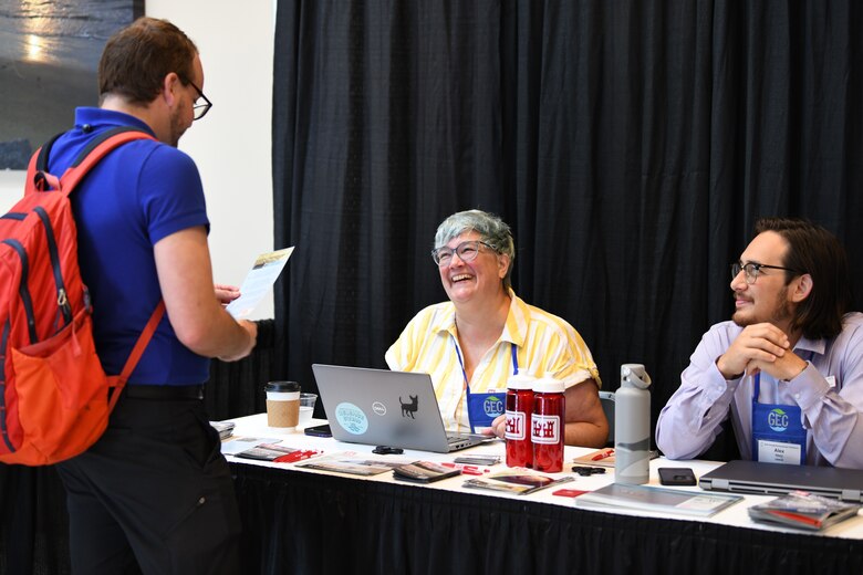 Photo of a male conference attendee standing in front of a table talking to two USACE employees.