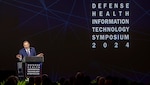 Man in suit speaks at podium in front of sign that says "DEFENSE HEALTH INFORMATION TECHNOLOGY SYMPOSIUM 2024."