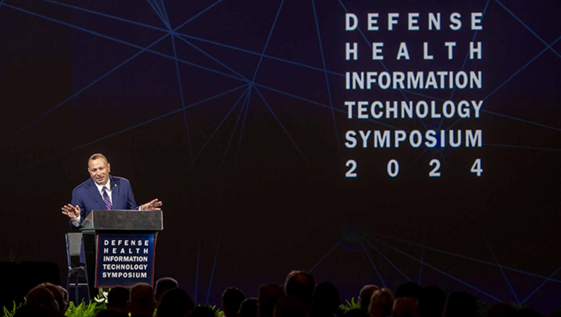 Man in suit speaks at podium in front of sign that says "DEFENSE HEALTH INFORMATION TECHNOLOGY SYMPOSIUM 2024."