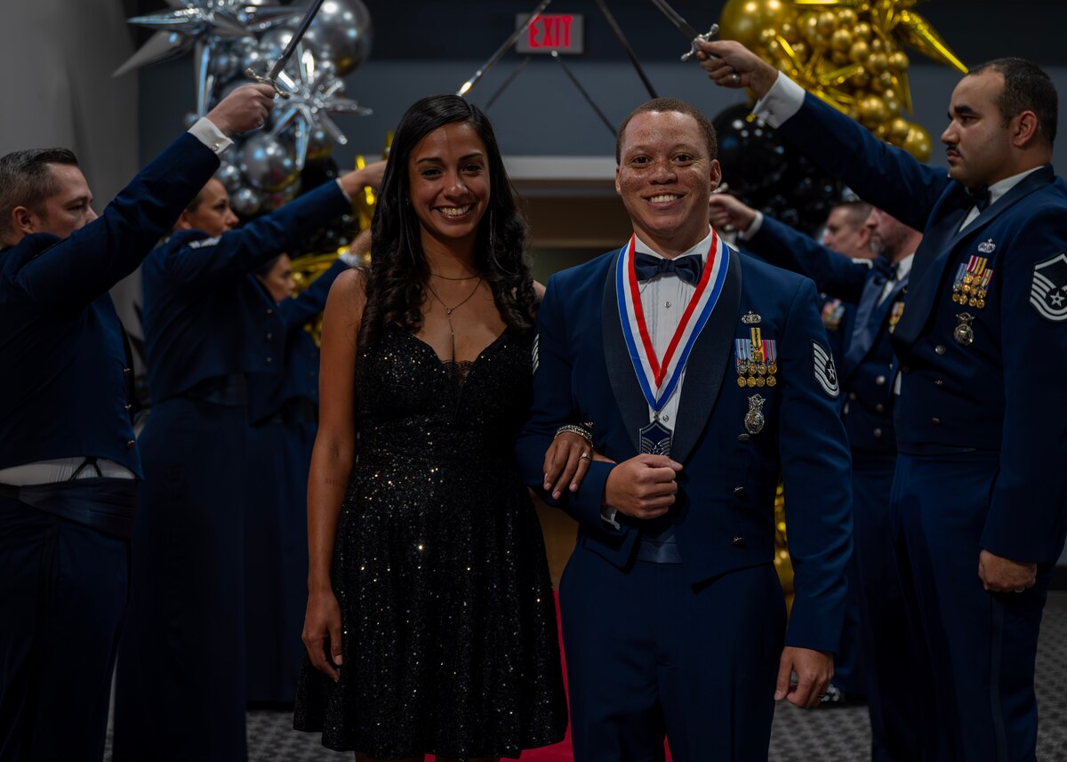 U.S. Air Force Master Sgt. Select Demarcus Hayes, 17th Security Forces Squadron operations noncommissioned officer, and his spouse, Mi Ami Hayes, walk through the saber cordon, inducting him into the highest tier of the enlisted force during the Senior Noncommissioned Officer Induction Ceremony at the Powell Event Center, Goodfellow Air Force Base, Texas, Aug. 23, 2024. Each inductee entered the room through a saber cordon, which is used to salute the new SNCOs to show respect for the sacrifice and commitment the inductees have taken to earn the rank of master sergeant. (U.S. Air Force photo by Senior Airman Sarah Williams)