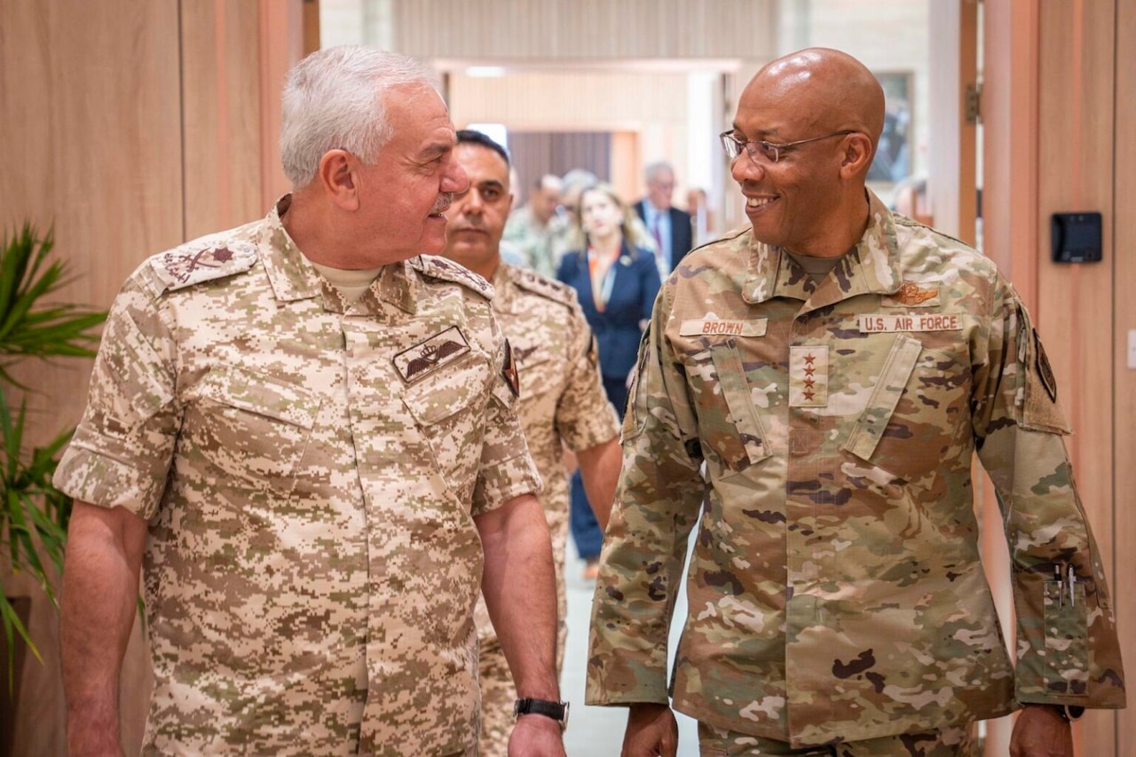 Two men in camouflage military uniforms are walking indoors toward the camera while talking to each other.