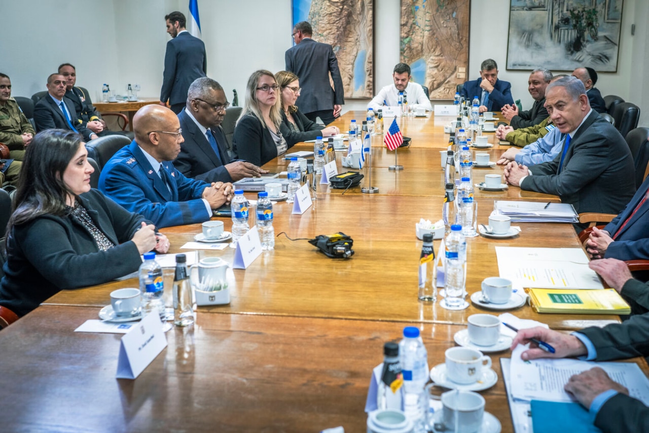 Roughly one dozen men and women, most of them in business attire and one of them in a dress military uniform, are sitting around a long wooden table in an office, having a conversation. Bottles of water, coffee cups and notes rest on the table in front of them.