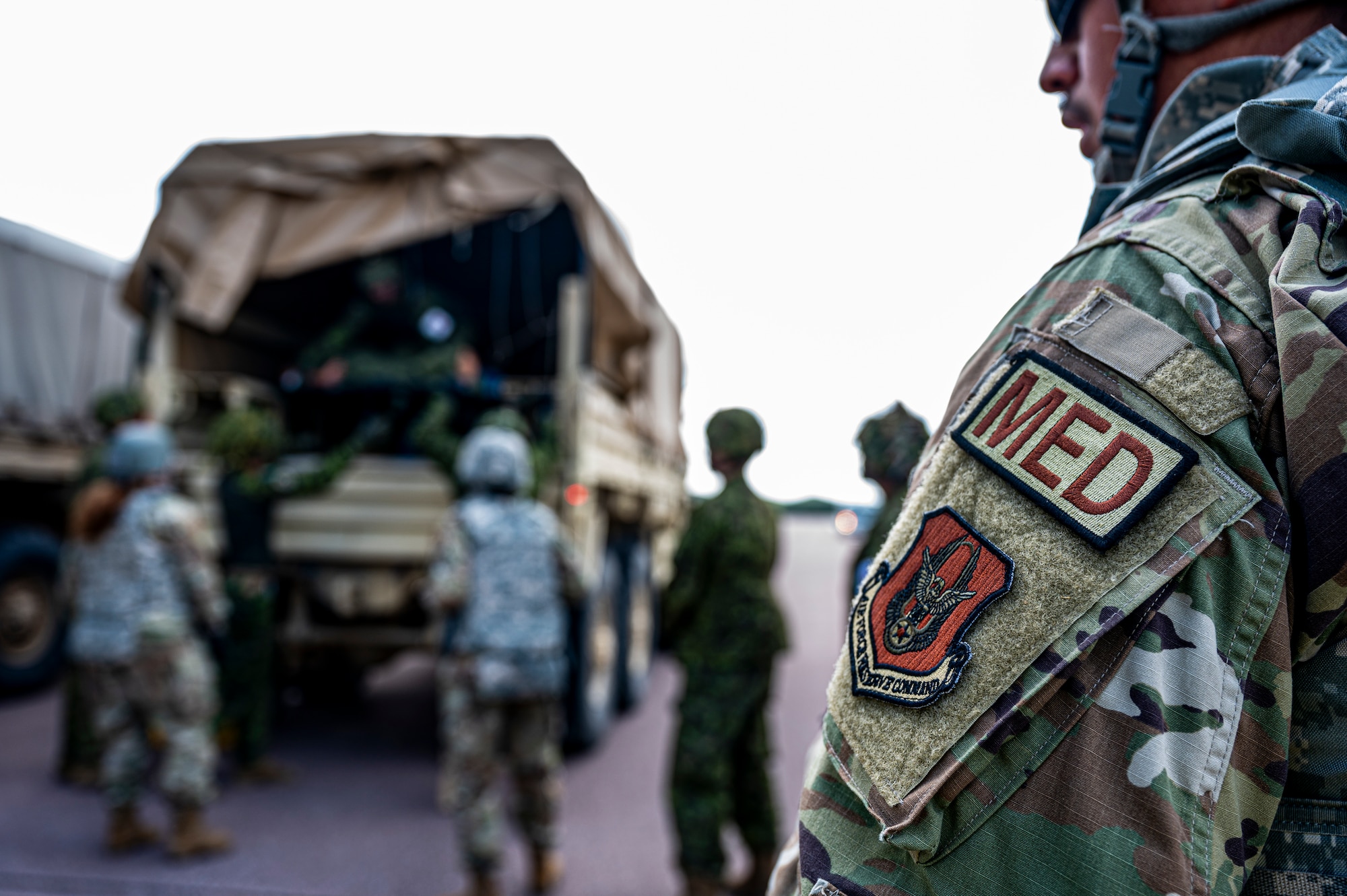 The duty identifier patch titled "MED" signifying medical personnel on the sleeve of an Air Force reservist sits in right side of frame as medics transfer patients in the background.
