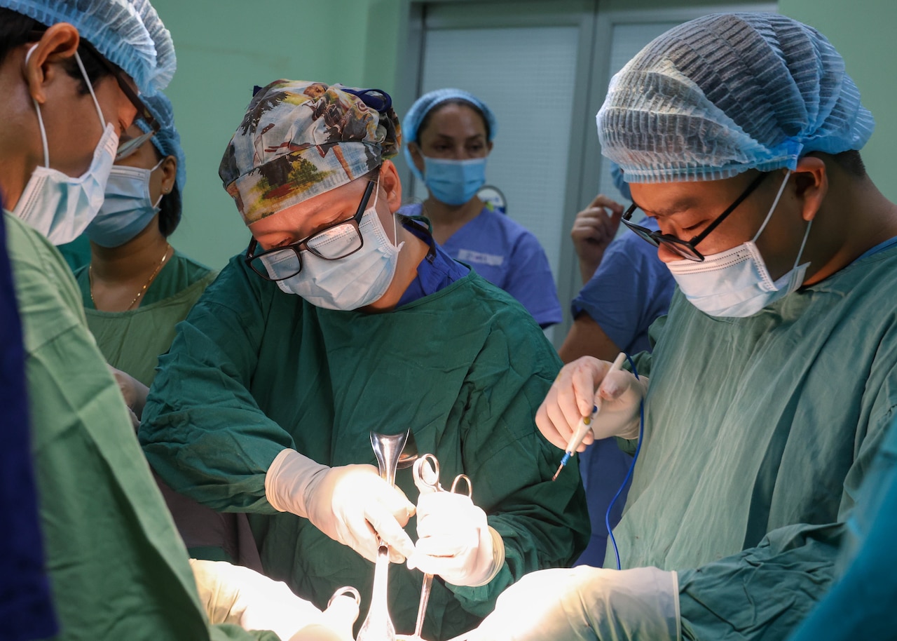 Two people in scrubs and masks hold surgical tools over a table as others surround them.