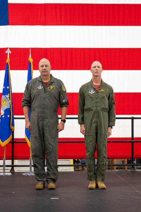 Two men in olive green flight suits stand at attention in front of a large American flag.