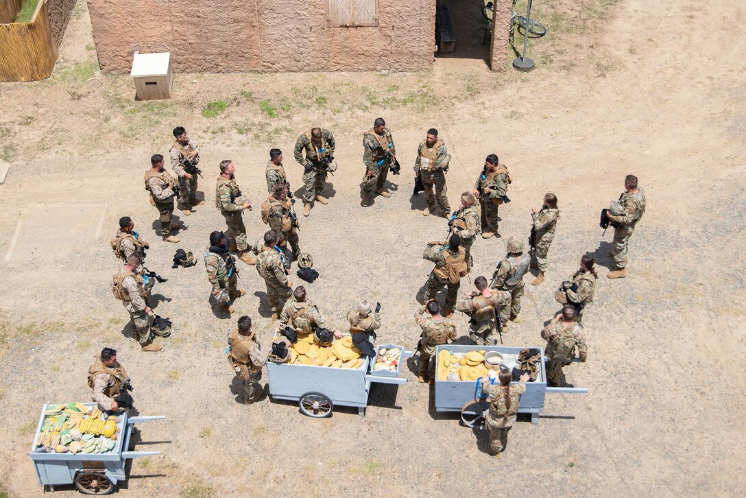 Dozens of service members gather in a large circle on a dirt road outside a concrete structure near three carriages carrying fruits and vegetables.