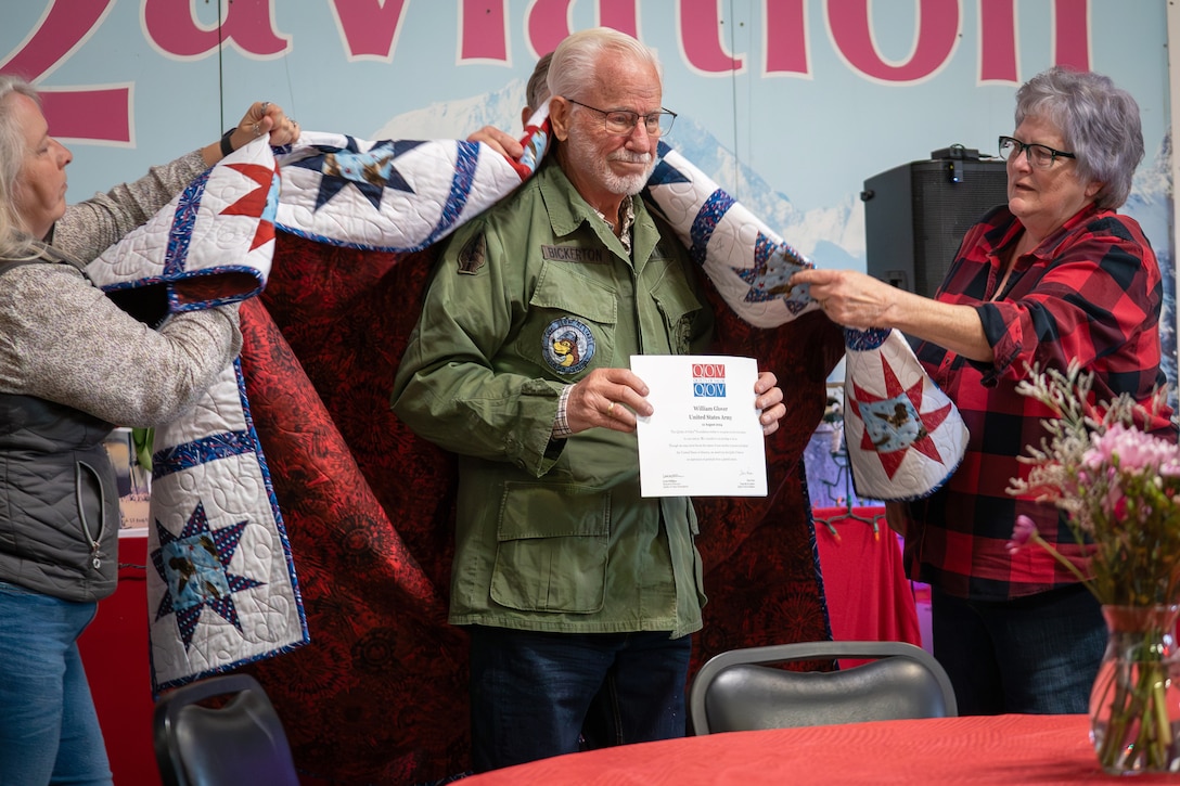 Three people put a quilt on the back of a veteran as they hold a sign.