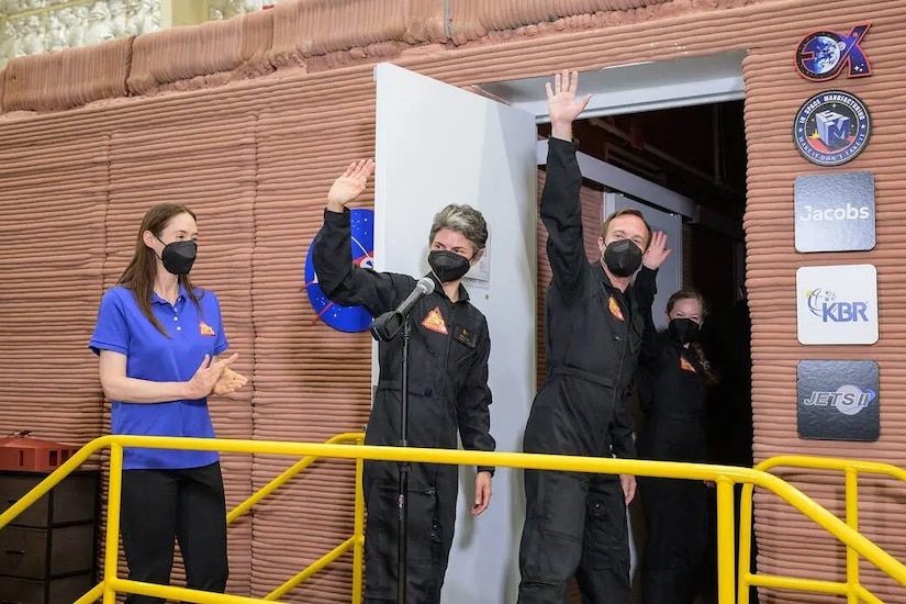 Three people wave to others as they walk into a building. A fourth person near them applauds.