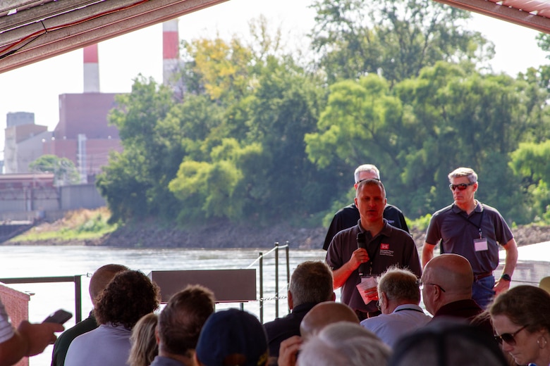 Three men in black shirts stand in front of a group of people sitting with a river and trees in the background.