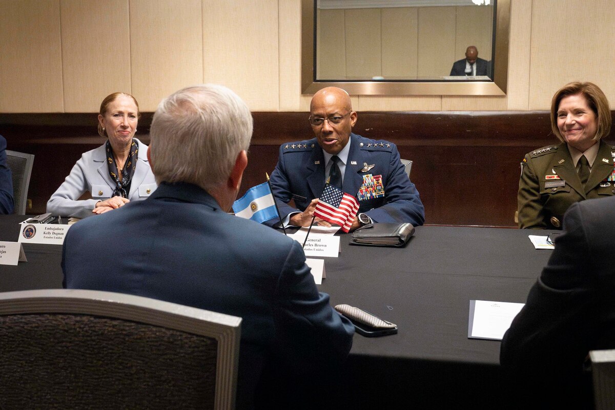 A woman in business attire, a man in a dress military uniform and a woman in a dress military uniform sit at a table in a conference room talking to a man on the other side of the table with his back to the camera.