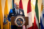 A man in military dress uniform stands at a lectern delivering remarks. Seven flags of various South American countries are behind him.