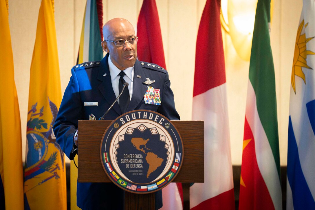 A man in military dress uniform stands at a lectern delivering remarks. Seven flags of various South American countries are behind him.
