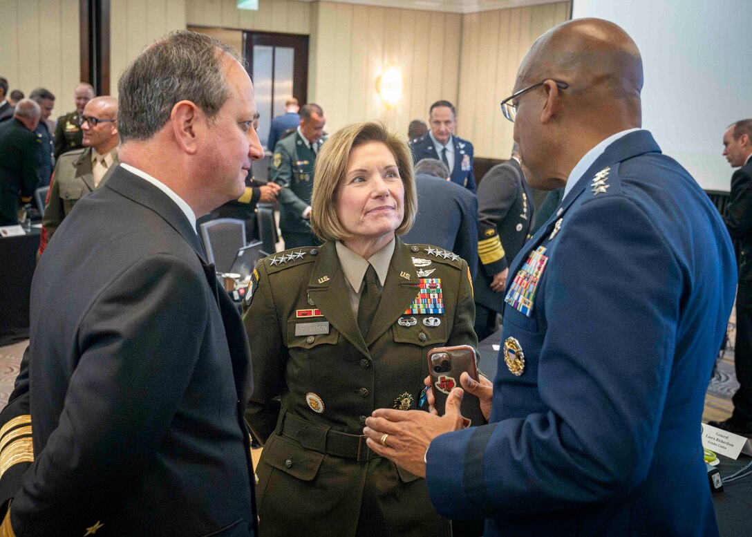 A man in business attire, a woman in dress military uniform and another man in dress military uniform stand while conversing in a conference hall.