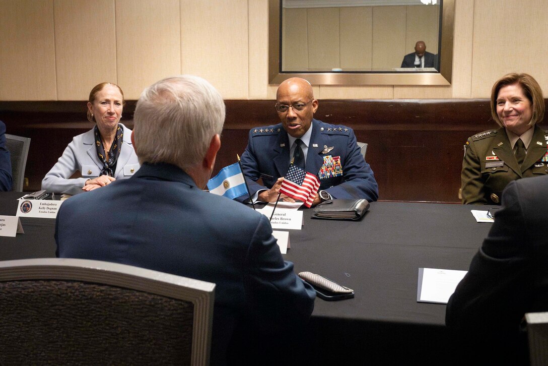 A woman in business attire, a man in a dress military uniform and a woman in a dress military uniform sit at a table in a conference room talking to a man on the other side of the table with his back to the camera.