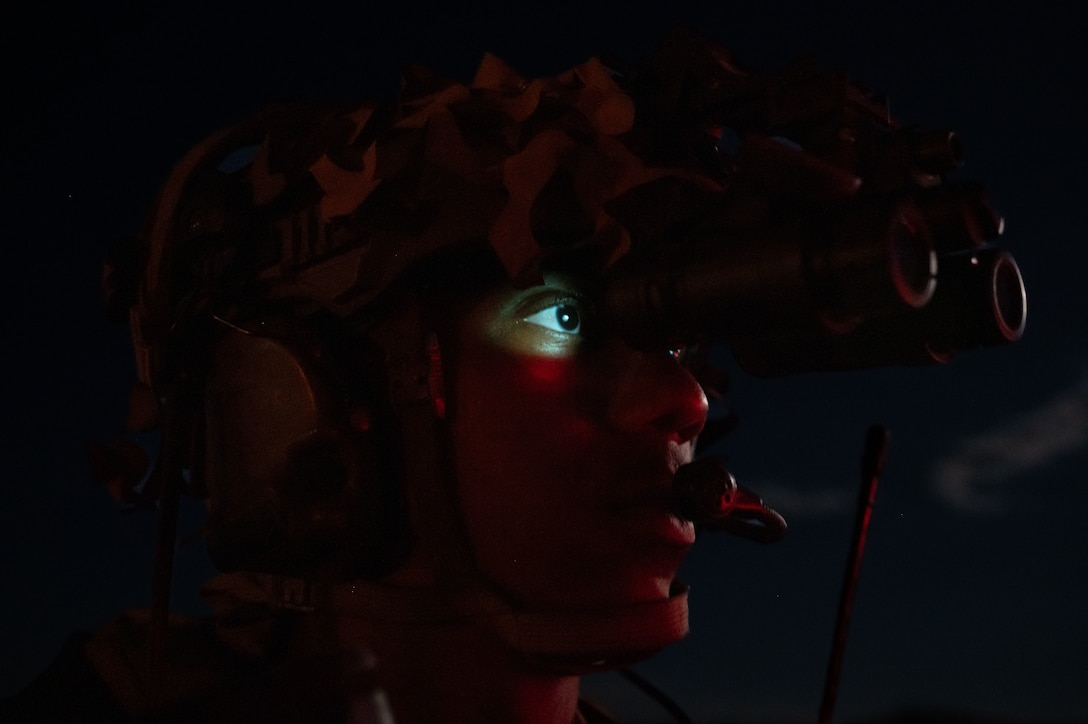 An airman wearing a helmet and combat gear looks through binoculars at night.