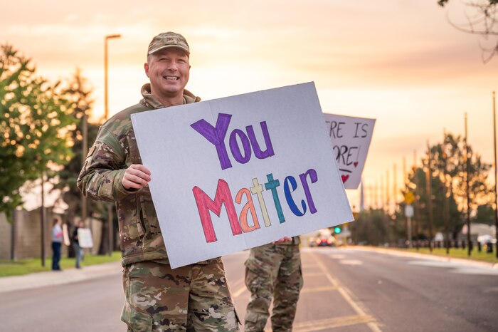 A man in camo uniform holds a white poster with words while standing in the middle of the street.