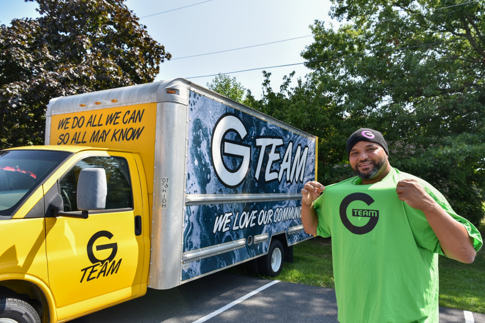 Man wearing beanie and green t-shirt that says "G TEAM" stands in front of yellow moving truck.