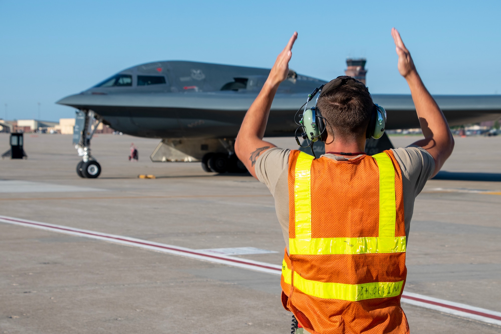 A U.S. Air Force Airman marshals a B-2 Spirit stealth bomber on the flight line at an Air Force Base. He is wearing a high-visibility orange and yellow safety vest, and signaling the aircraft with his arms.