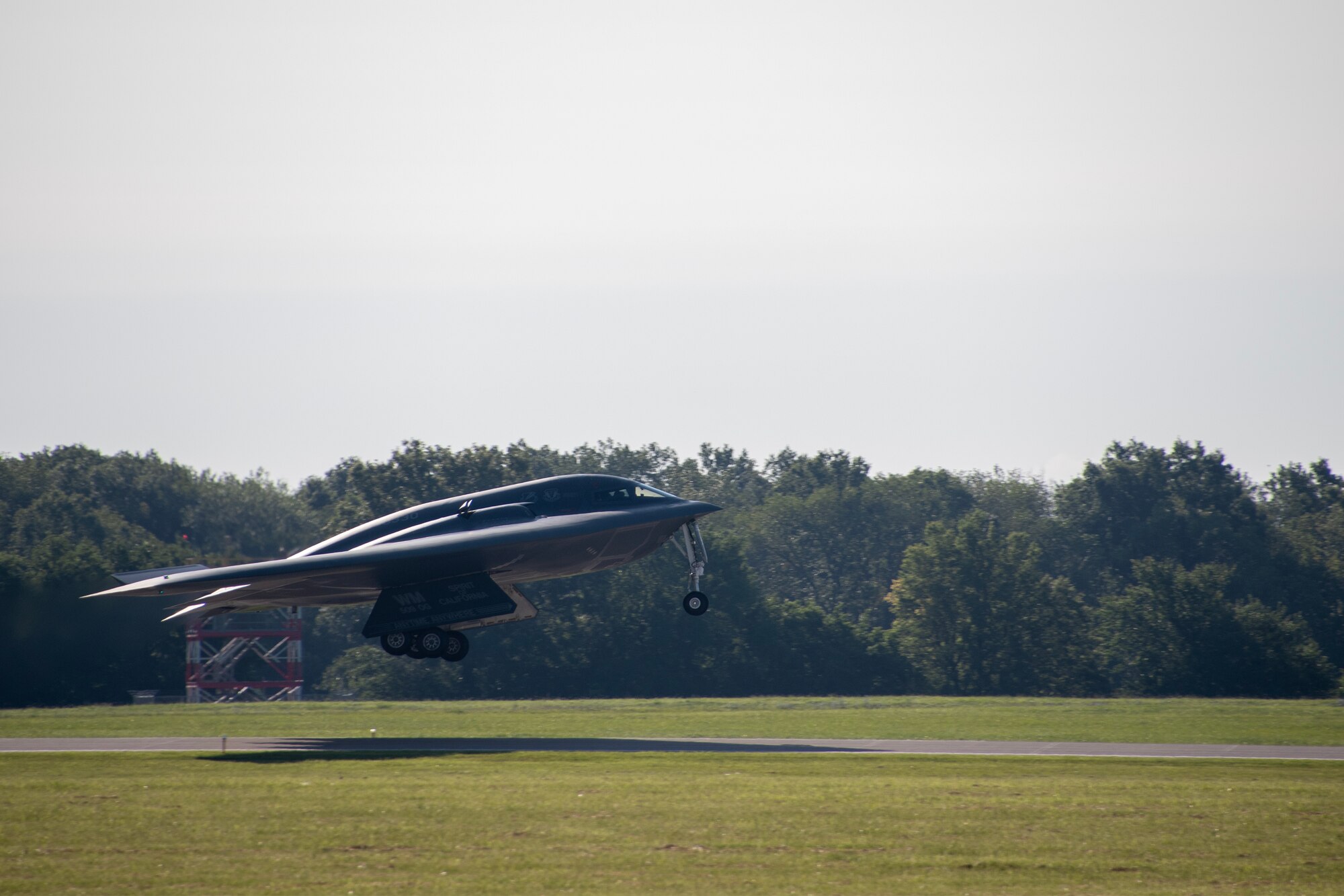A B-2 Spirit stealth bomber takes off from a runway. The runway is flanked by green grass and trees.