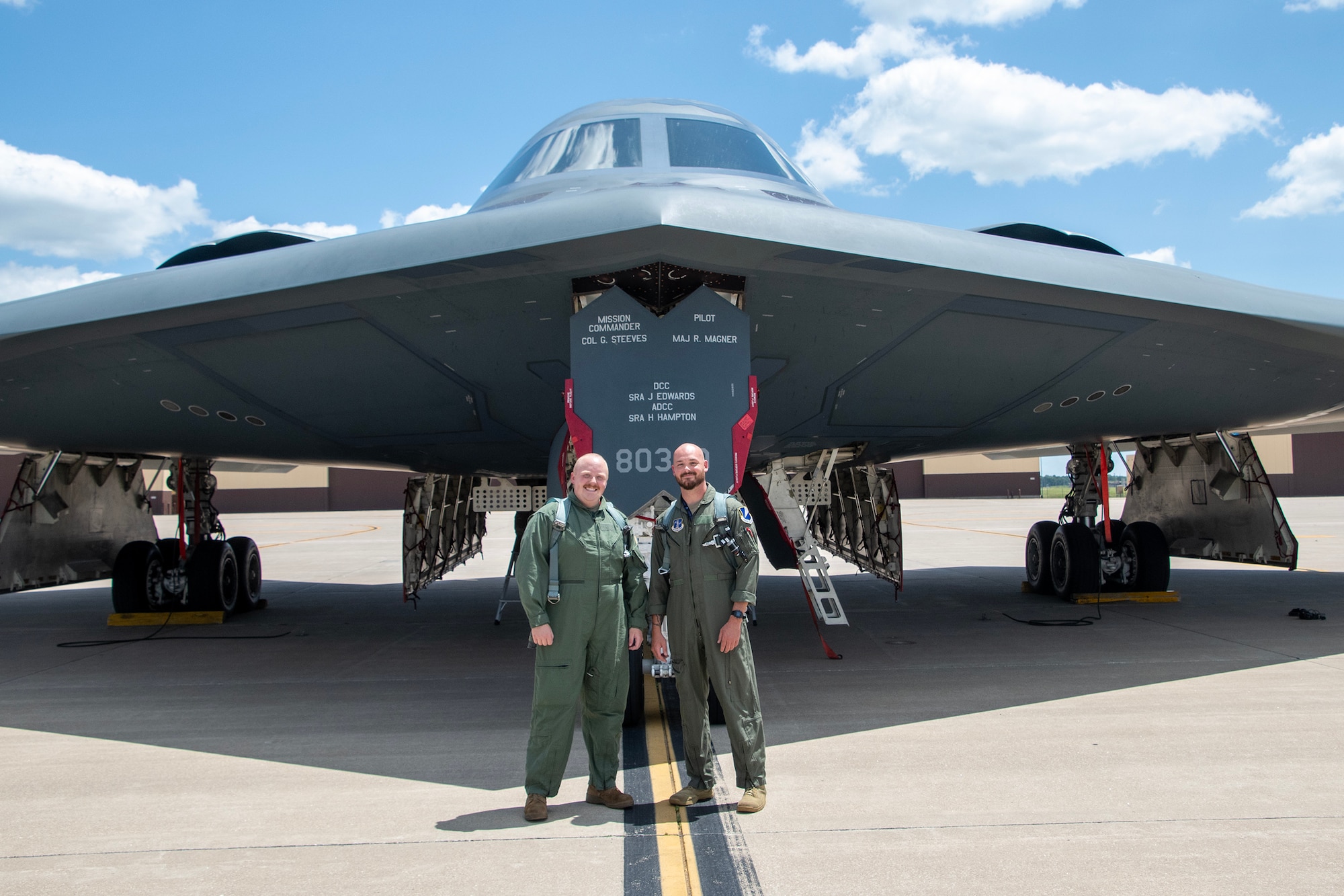 Two U.S. Air National Guard Airmen stand in front of a B-2 Spirit stealth bomber on a flight line. The Airmen are wearing flight suits.