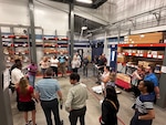 A group of men and women stand listening to a speaker in a warehouse