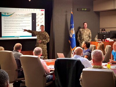 A white man in camo uniform points to a large screen while talking to men and women sitting at a table