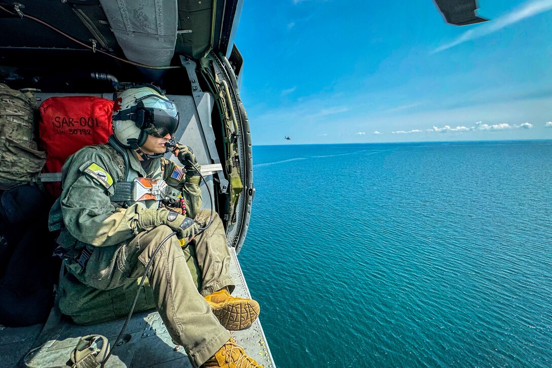 A sailor wearing a helmet sits in the open doorway of a helicopter as it flies over a body of water and another helicopter flies in the distance.