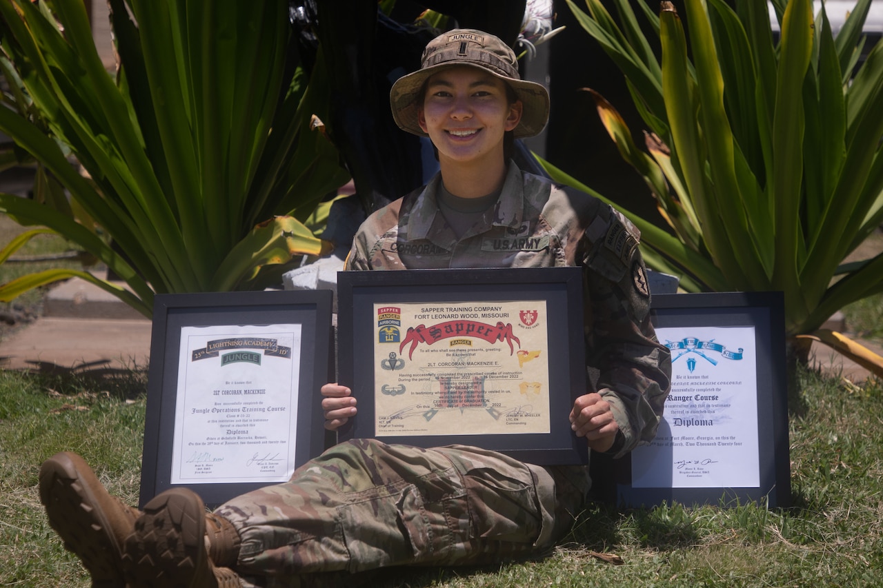 A soldier sits on the grass during a sunny day holding three certificates.