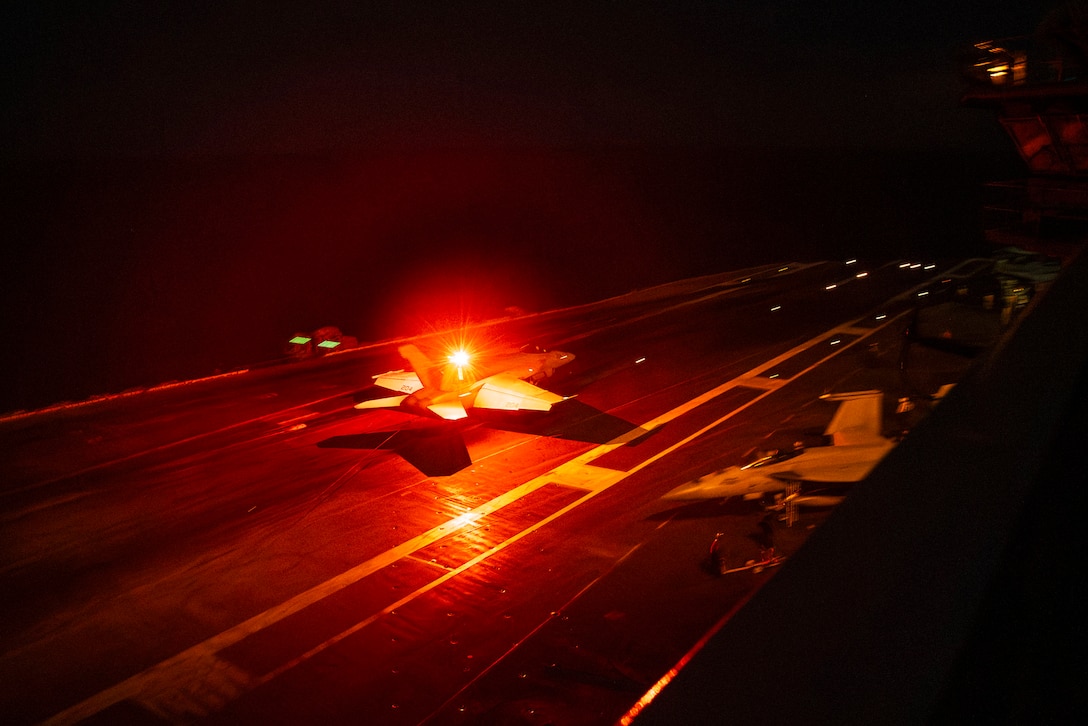 A military helicopter with a red light atop it lands on the flight deck of a Navy ship at night.