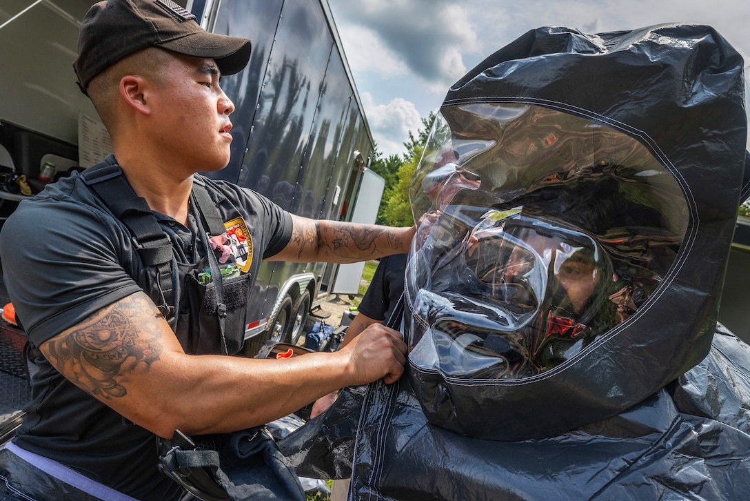 A service member adjusts the head covering portion of a protective suit for another service member during daylight.