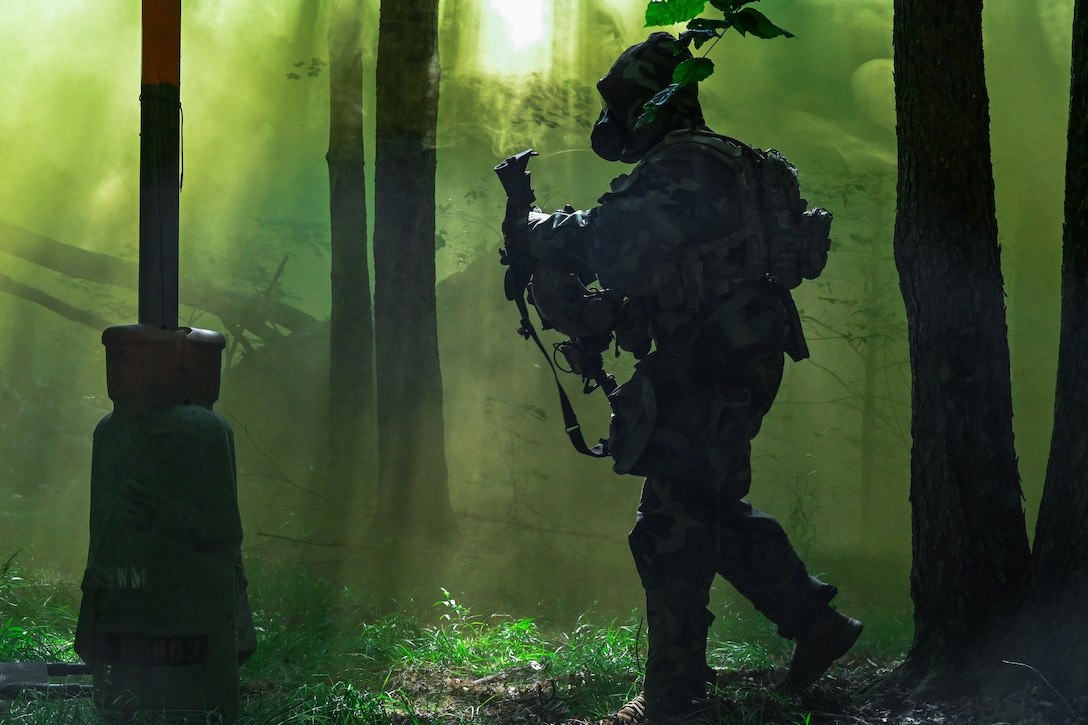 An airman wearing protective gear walks through a forest carrying a weapon surrounded by a haze of yellow and green smoke.