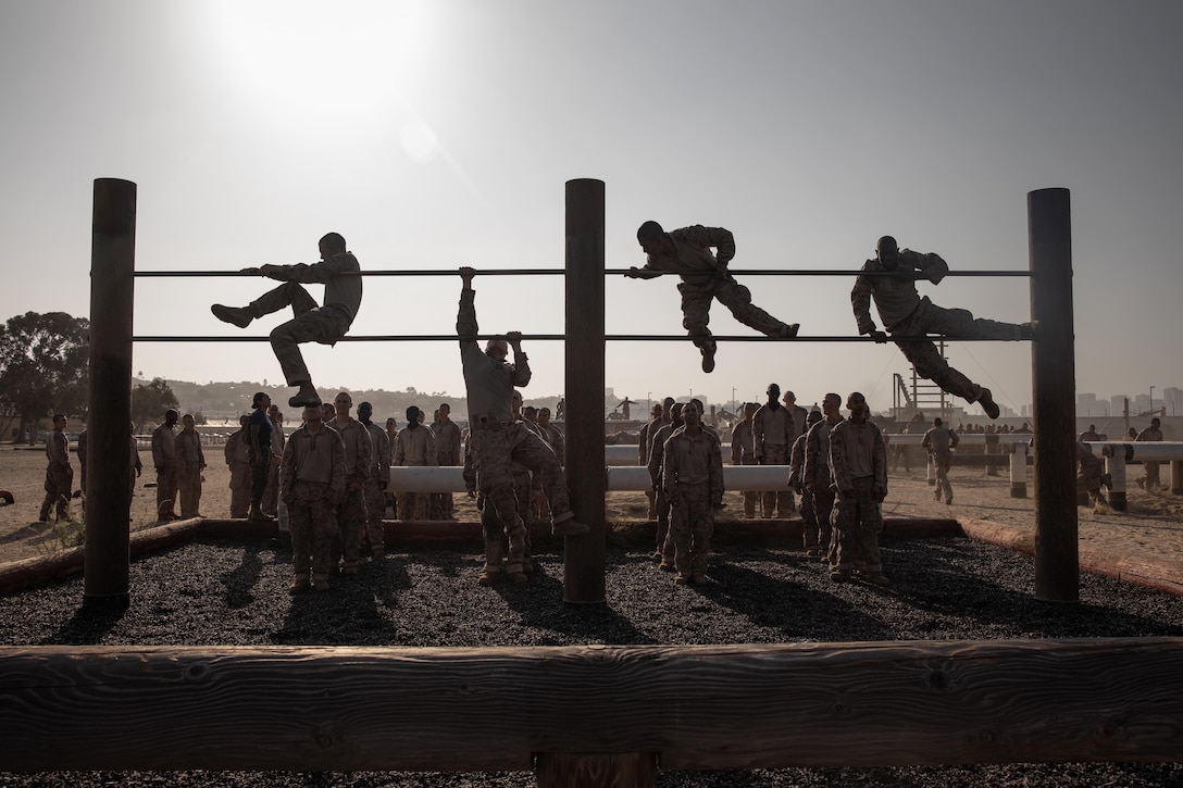 Four Marine Corps recruits climb a jungle gym as fellow recruits watch while standing in formation under a sunlit sky as seen in silhouette.