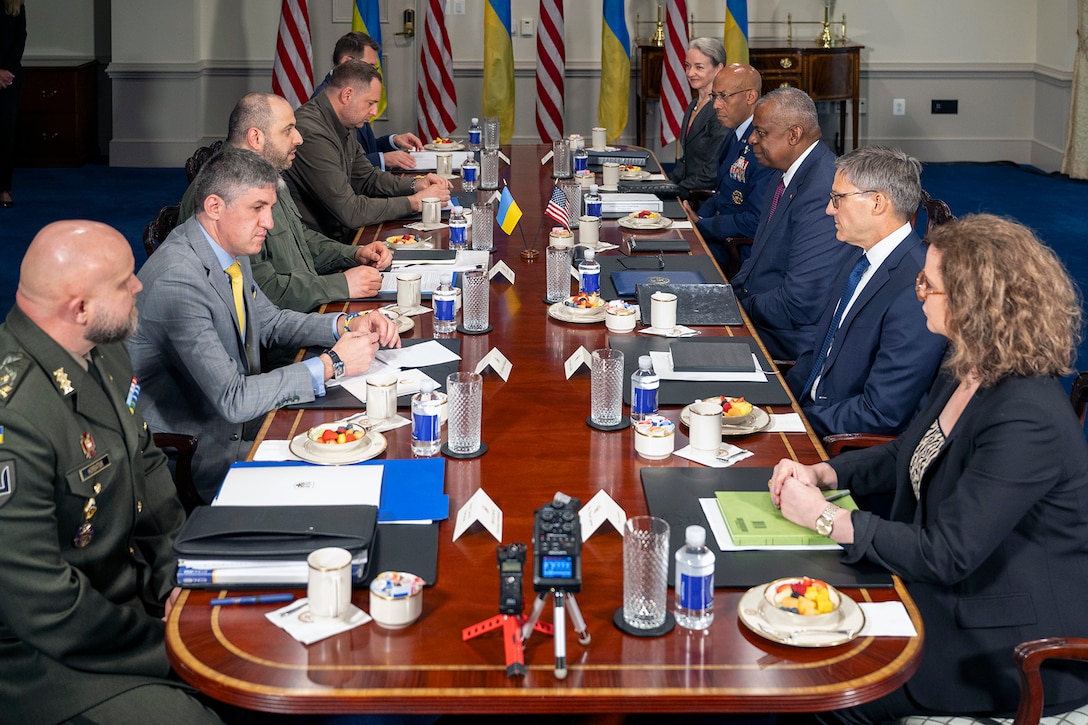 A group of government officials sit around a long rectangular table by the Ukrainian and U.S. flags.