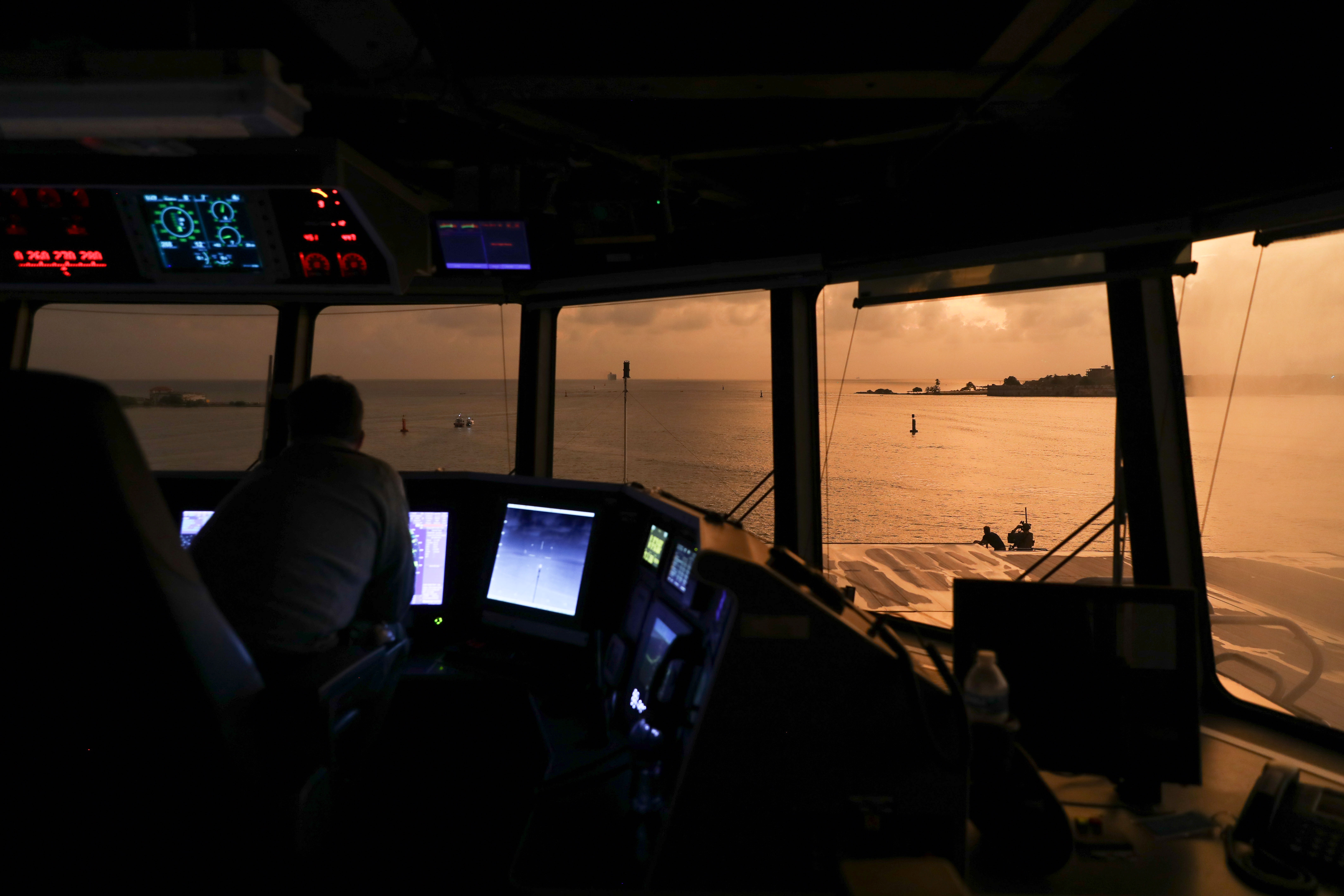 A service member sitting in a ship’s control room illuminated by colorful lights looks through the windshield at the ocean as sun shines through.