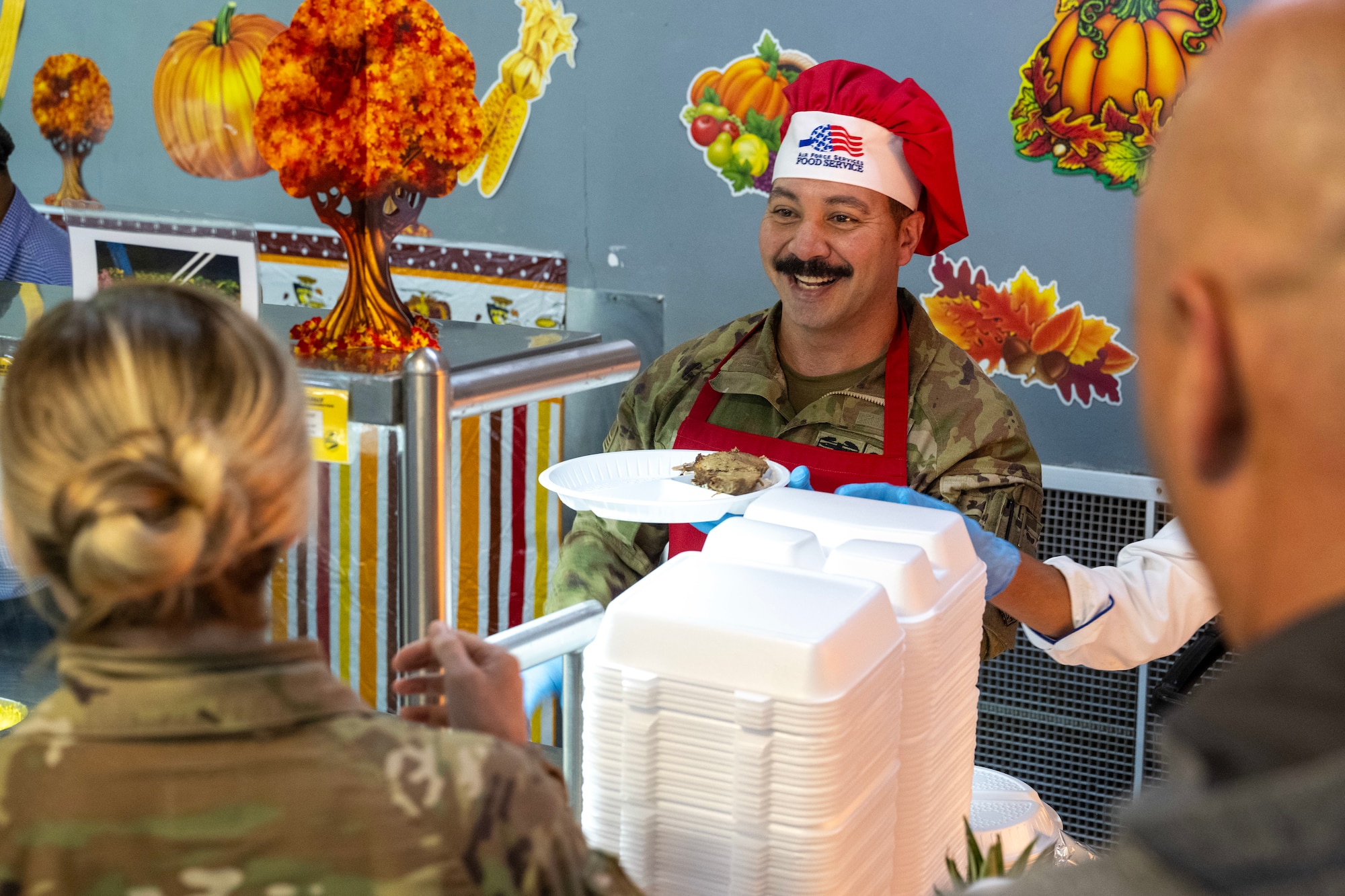 A chief master sergeant smiles as he serves a Thanksgiving meal to dining facility patrons.